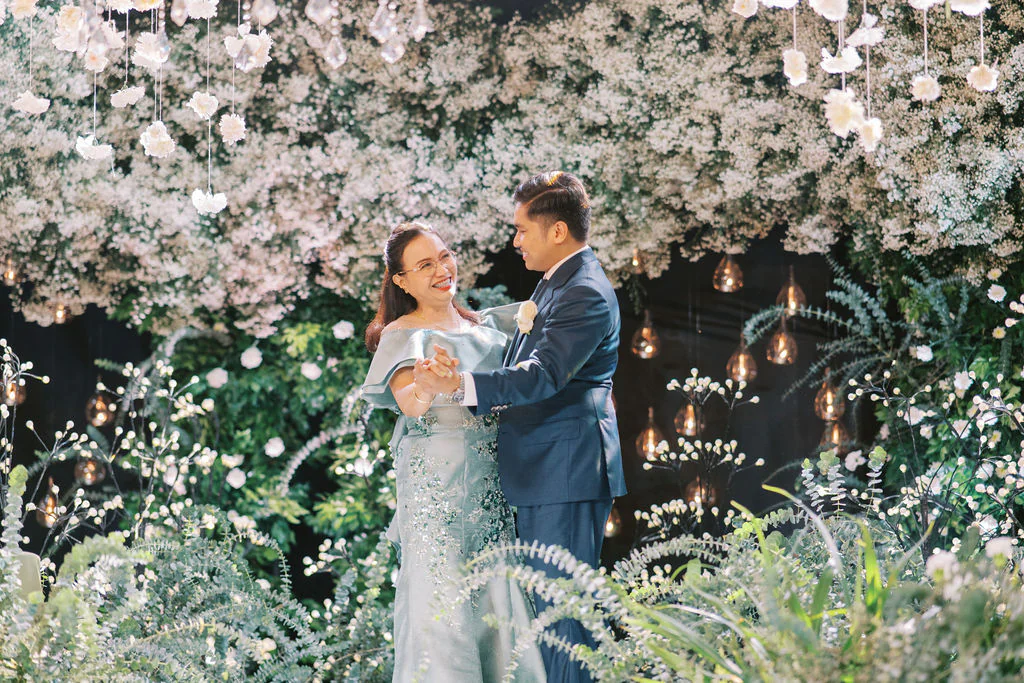 The groom and his mother share a dance in front of a backdrop of white florals and glowing candles