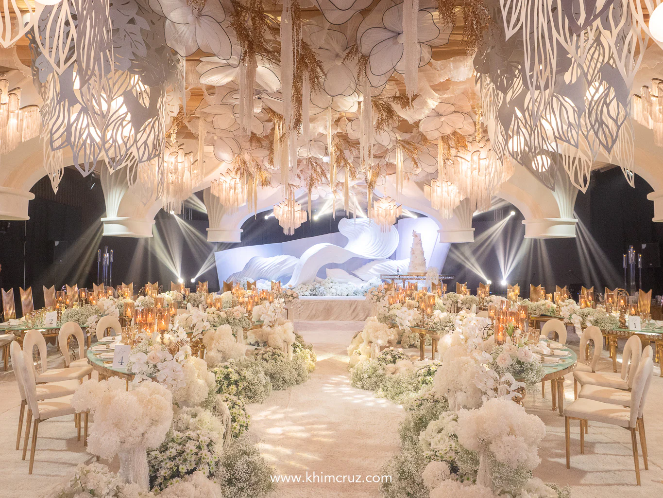 a wave-like backdrop sets the stage with suspended florals and arching ceiling structures add depth and elegance to this wedding reception